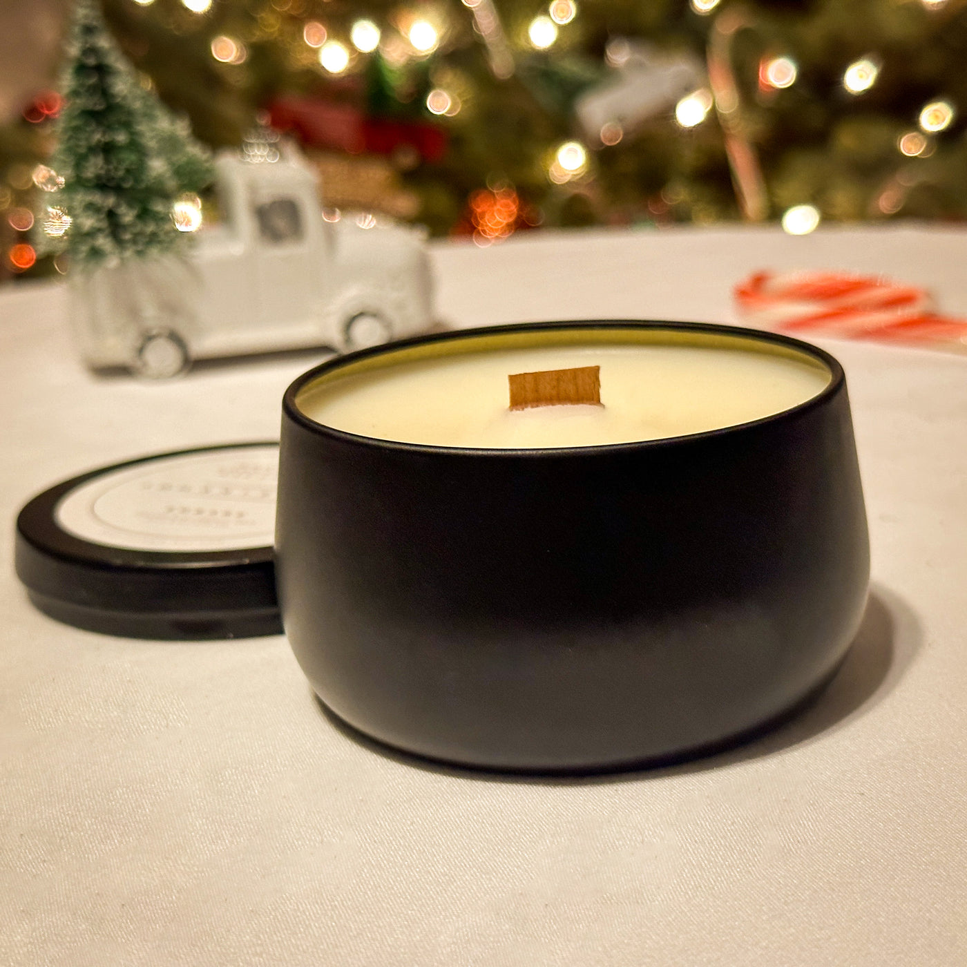 Black metal candle tin with white wax and wood wick, on a white table with a blurred Christmas scene in background