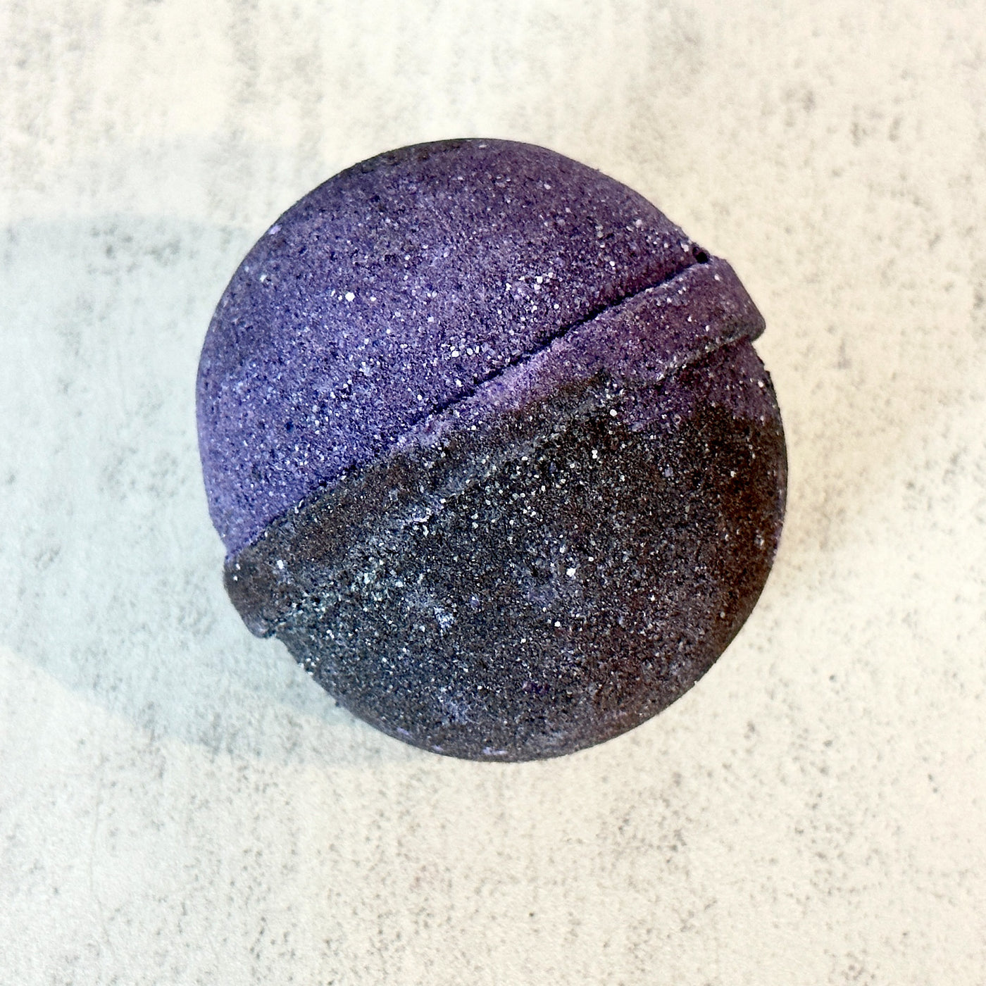 Purple and black Johnnie Ray Onyx bath bomb against concrete background.