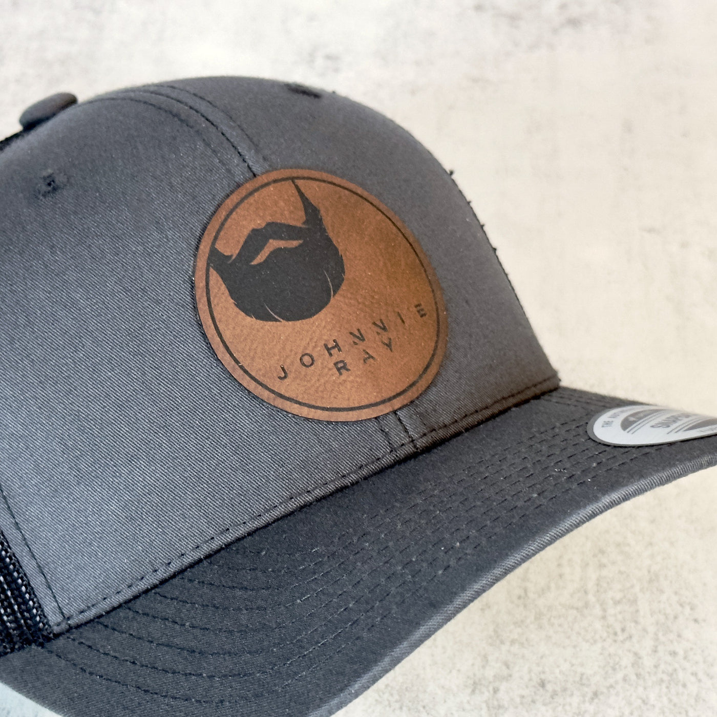 Angled view of Dark Grey Johnnie Ray hat with round brown leather patch with Johnnie Ray logo.