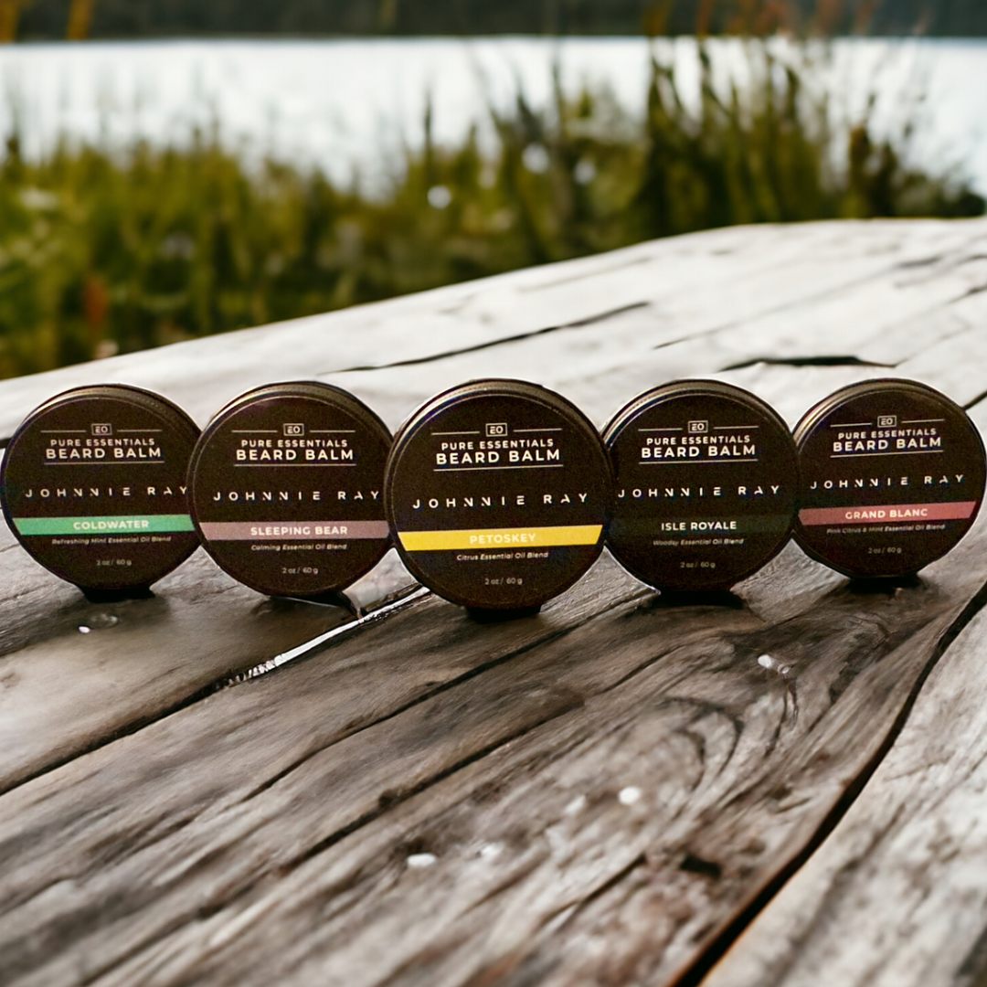 5 tins of Johnnie Ray Pure Essentials Beard Balm on rustic wood in front of grassy lake