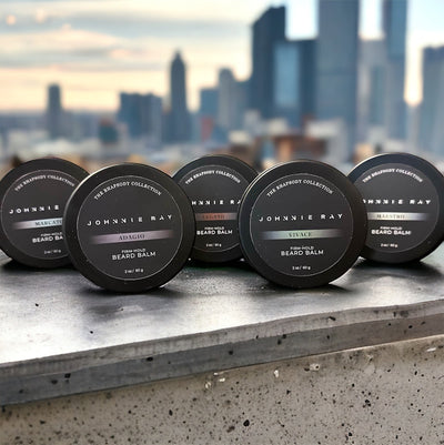 Five Johnnie Ray Beard Balms from The Rhapsody Collection in a black tin displayed on a concrete ledge overlooking city skyline