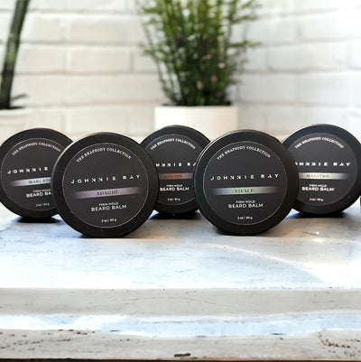 Five Johnnie Ray Beard Balms from The Rhapsody Collection in a black tin displayed on white granite countertop